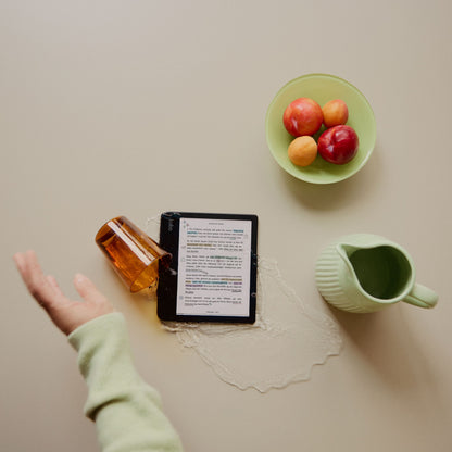 Kobo Libra Colour læseplade 7" Sort på bord, vand spildt fra væltet ravfarvet glas, grøn kande og frugtskål ved siden.