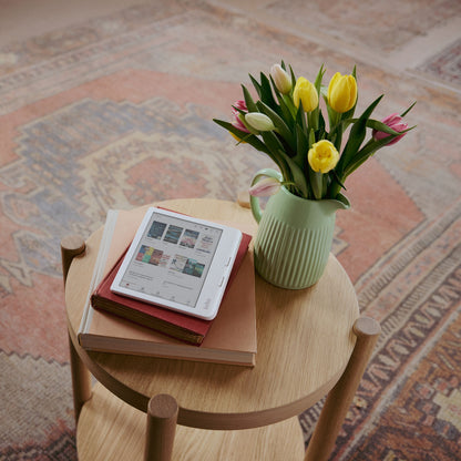 Kobo Libra Colour læseplade 7" Sort viser bogforsider, placeret på bord med bøger og mintgrøn kande med tulipaner.