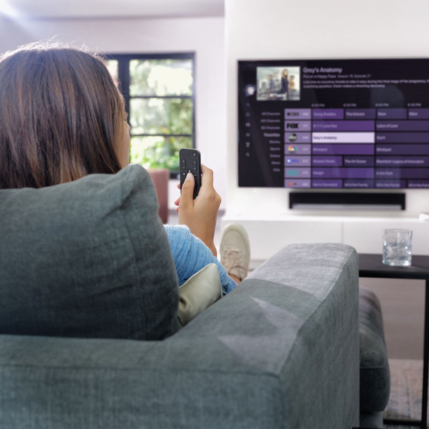 Apple TV-fjernbetjening En til alle sorter ses i hånd foran tv med programguide, grå sofa og glas på bord tæt ved.