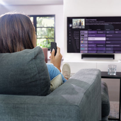 Apple TV-fjernbetjening En til alle sorter ses i hånd foran tv med programguide, grå sofa og glas på bord tæt ved.