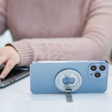 IPhone 12 blå med magnetisk mobilholder og flaskeåbner på bord, person i pink sweater skriver på laptop bagved.