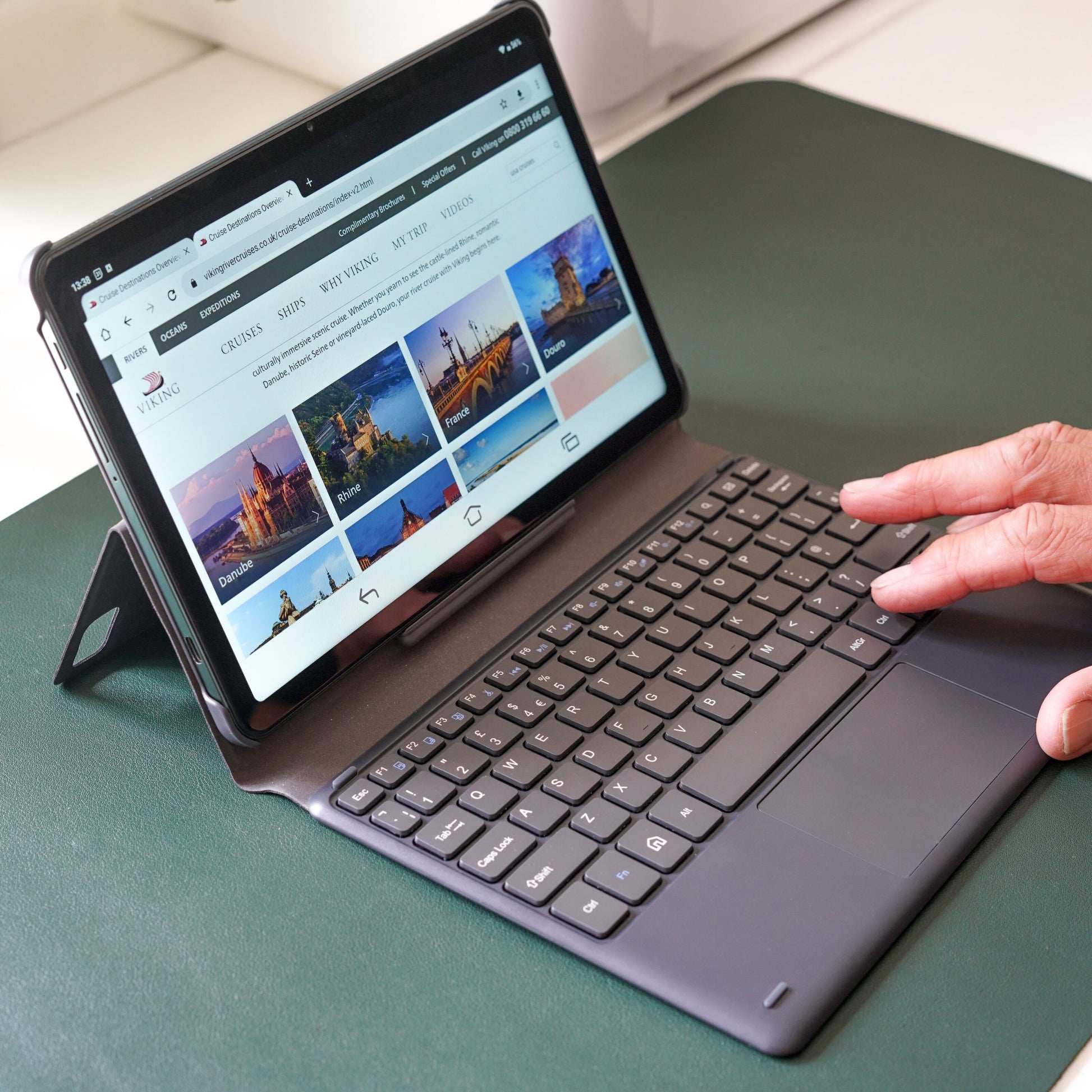 Doro Tablet med Doro Tastaturcover, vises på grøn skrivebordsunderlag, skærm viser rejsedestinationer og billeder.