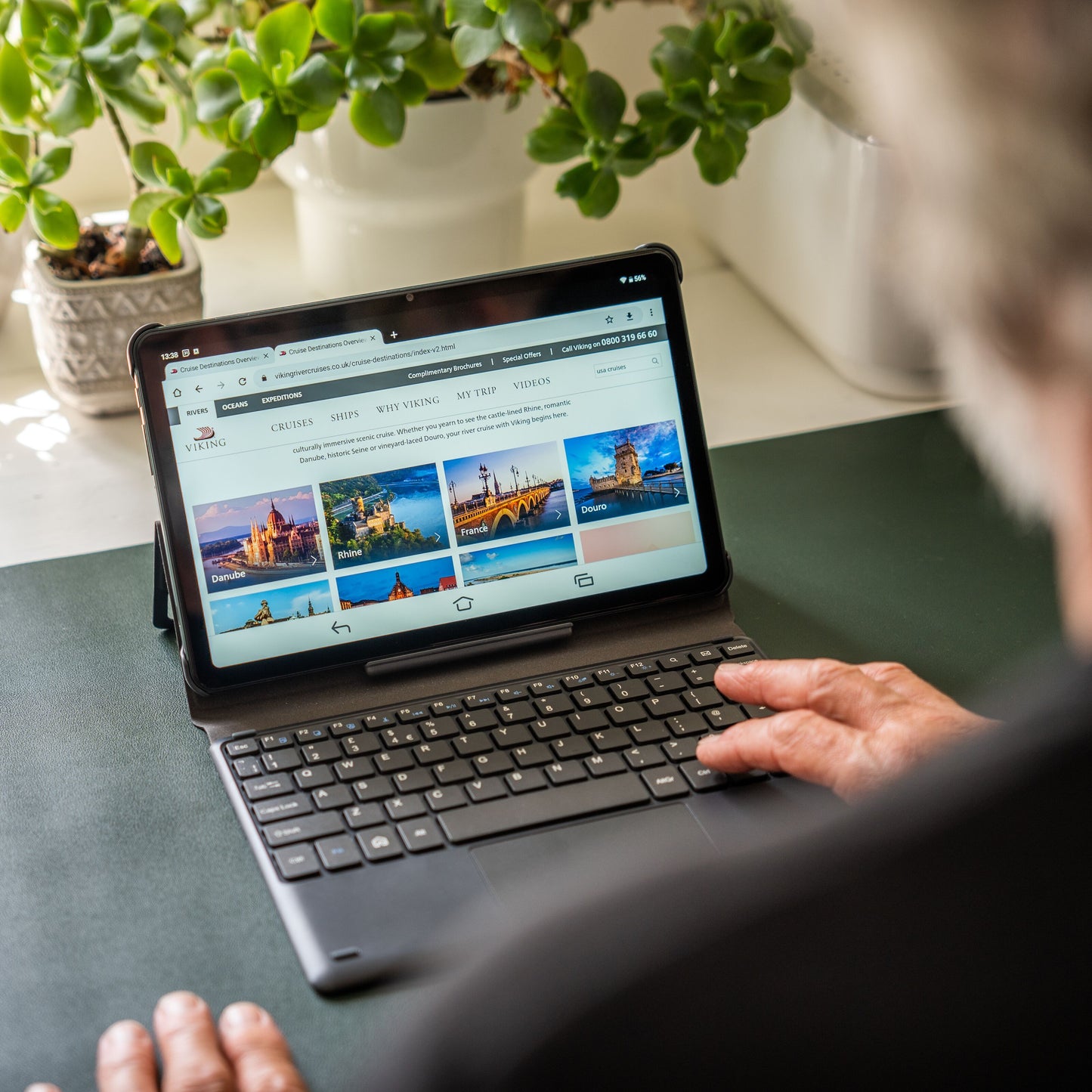 Doro Tastaturcover til Doro Tablet i brug, synlig skærm med rejsewebsite, grøn stueplante bagved.