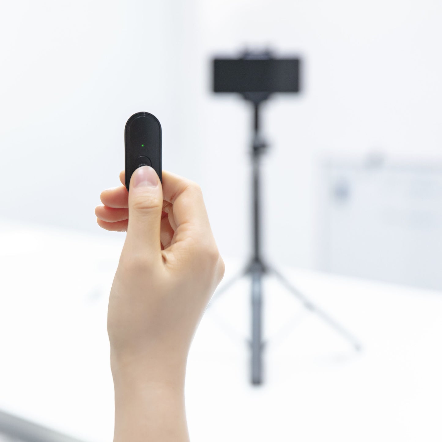 Linocell Bluetooth-udløser sort med grøn LED, holdes i hånden, sløret smartphone på Linocell Selfiestang Pro i baggrunden.