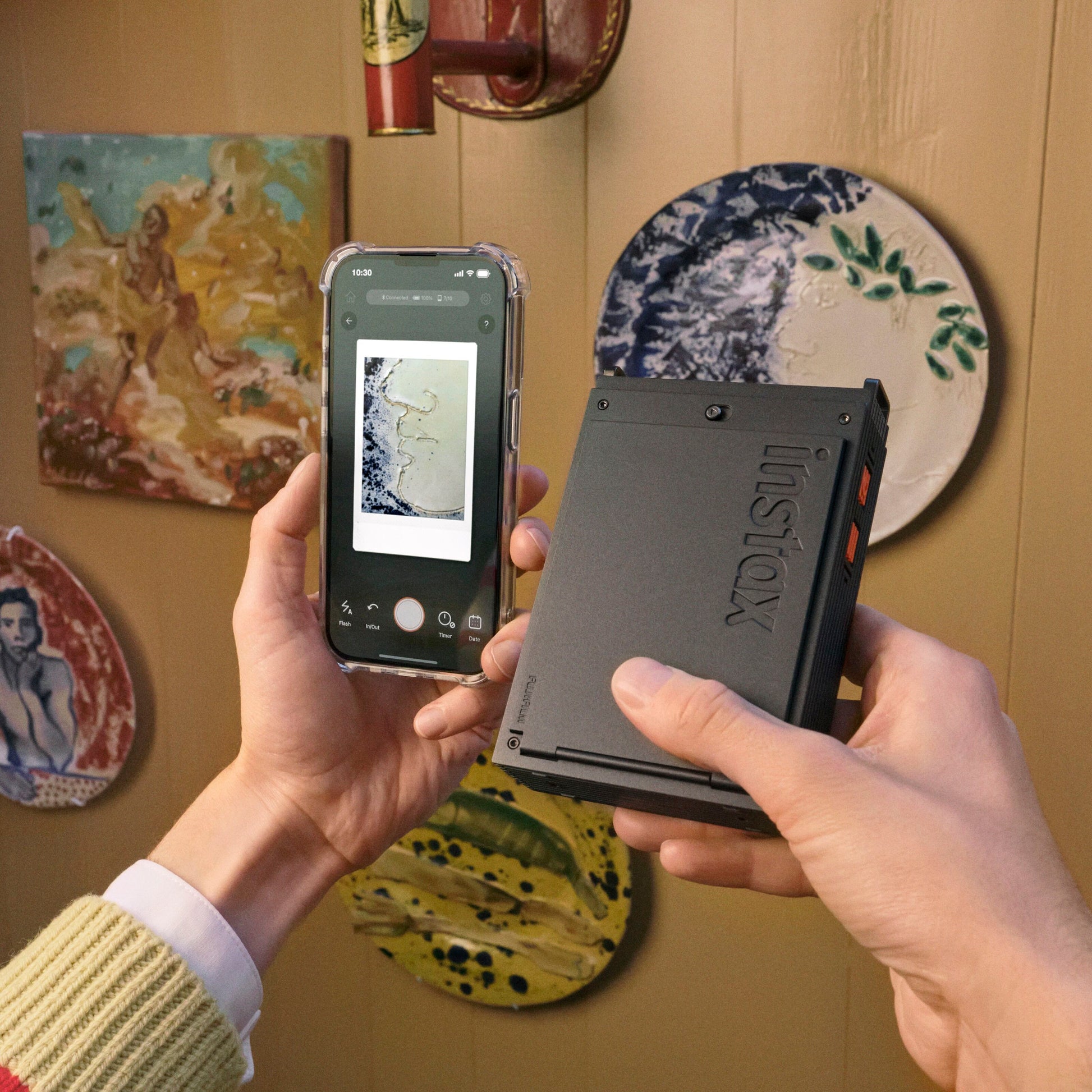 Sort Fujifilm instax mini Link+ mobil fotoskriver holdes i hånden, smartphone ved siden af, farverige keramikplader i baggrund.