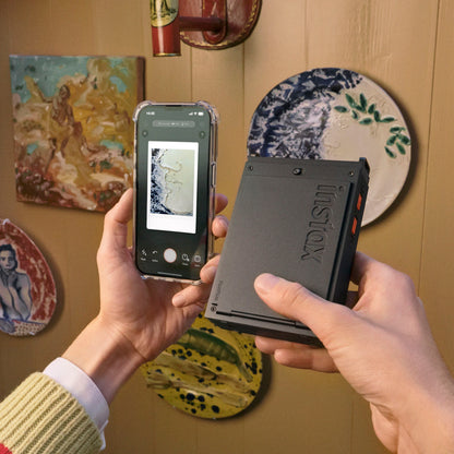 Sort Fujifilm instax mini Link+ mobil fotoskriver holdes i hånden, smartphone ved siden af, farverige keramikplader i baggrund.