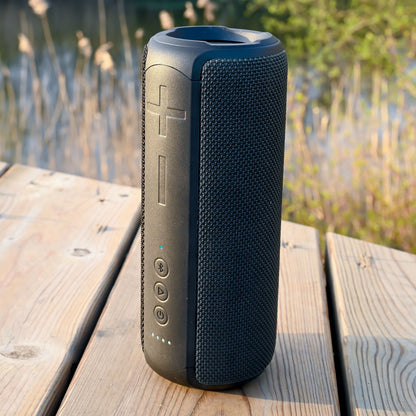 Nomadelic Loud 707 trådløs højttaler sort med store volumeknapper, Bluetooth 5.0 og IPX7, placeret på udendørs træbord.