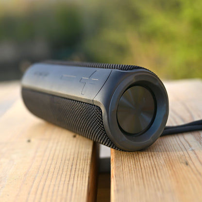 Nomadelic Loud 707 sort trådløs højttaler med Bluetooth 5.0 og IPX7 står på træoverflade, grønne planter bagved.