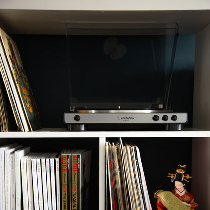 Audio Technica AT-LP60XUSB vinylafspiller med lukket låg på hylde over LP-plader og magasiner, dukke ses på nederste hylde.