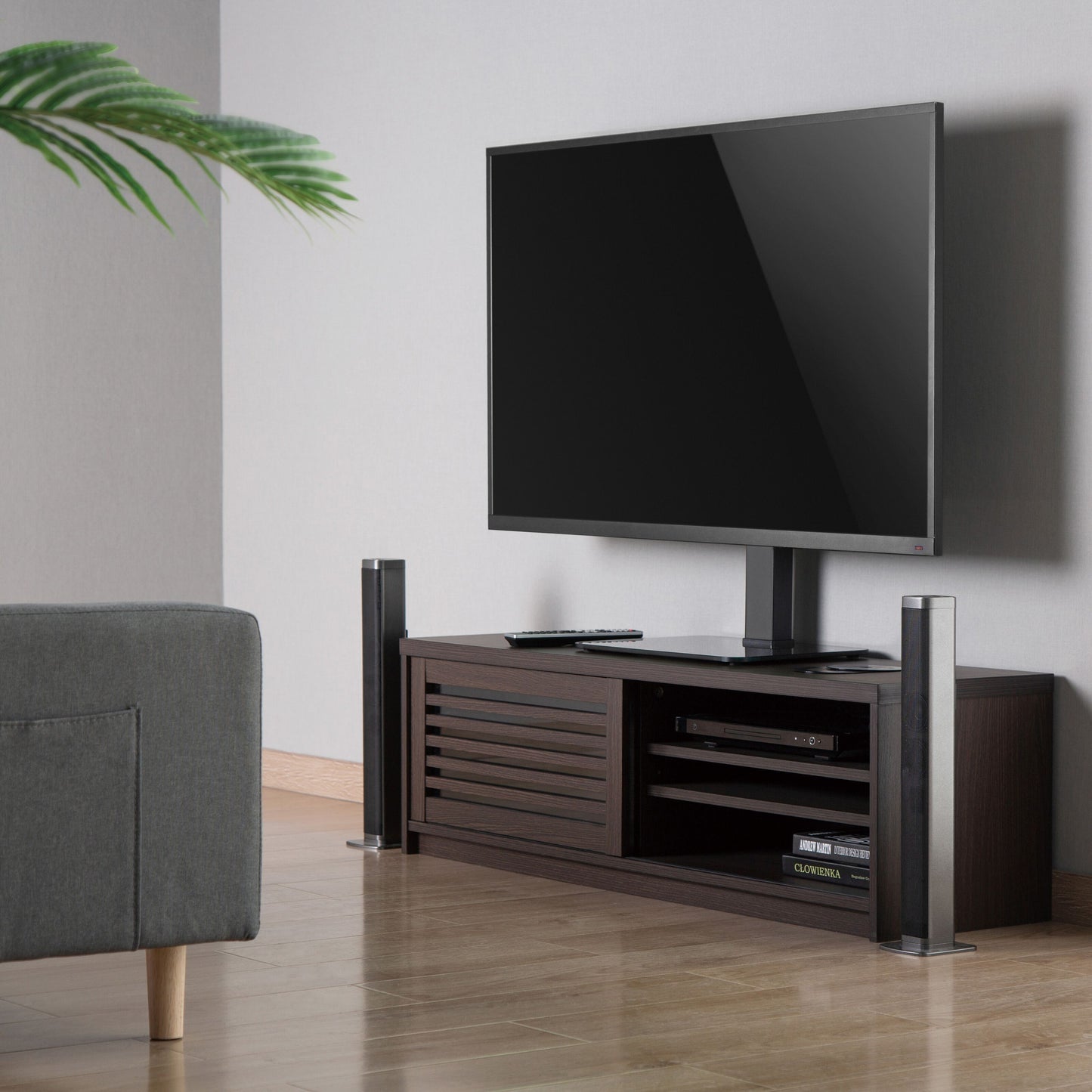 Fladskærms-TV på Luxorparts Bordstativ til TV 37-70" placeret på mørkt træbord med hylder, delvis grå sofa og grøn plante.