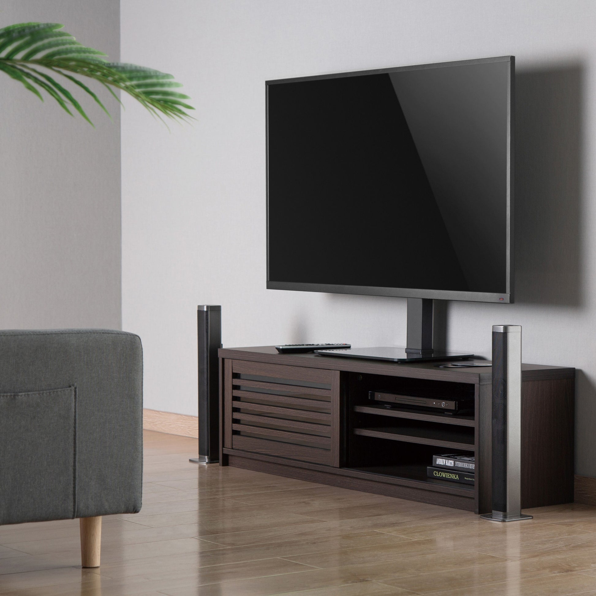 Fladskærms-TV på Luxorparts Bordstativ til TV 37-70" placeret på mørkt træbord med hylder, delvis grå sofa og grøn plante.