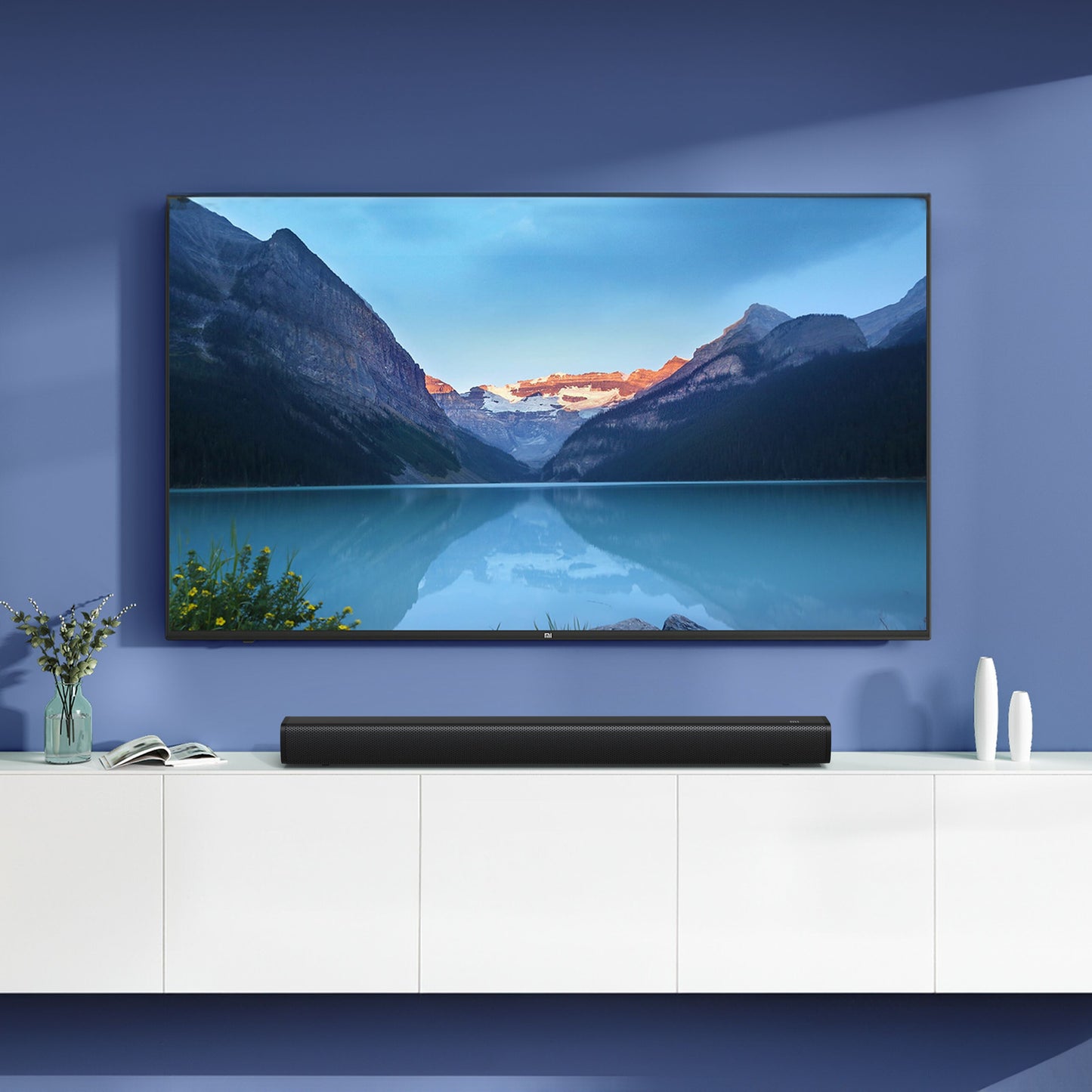 Xiaomi Soundbar 2.0 30W sort under vægmonteret TV med bjergsø-billede, hvidt skab, blomster og hvide objekter ved siden af.