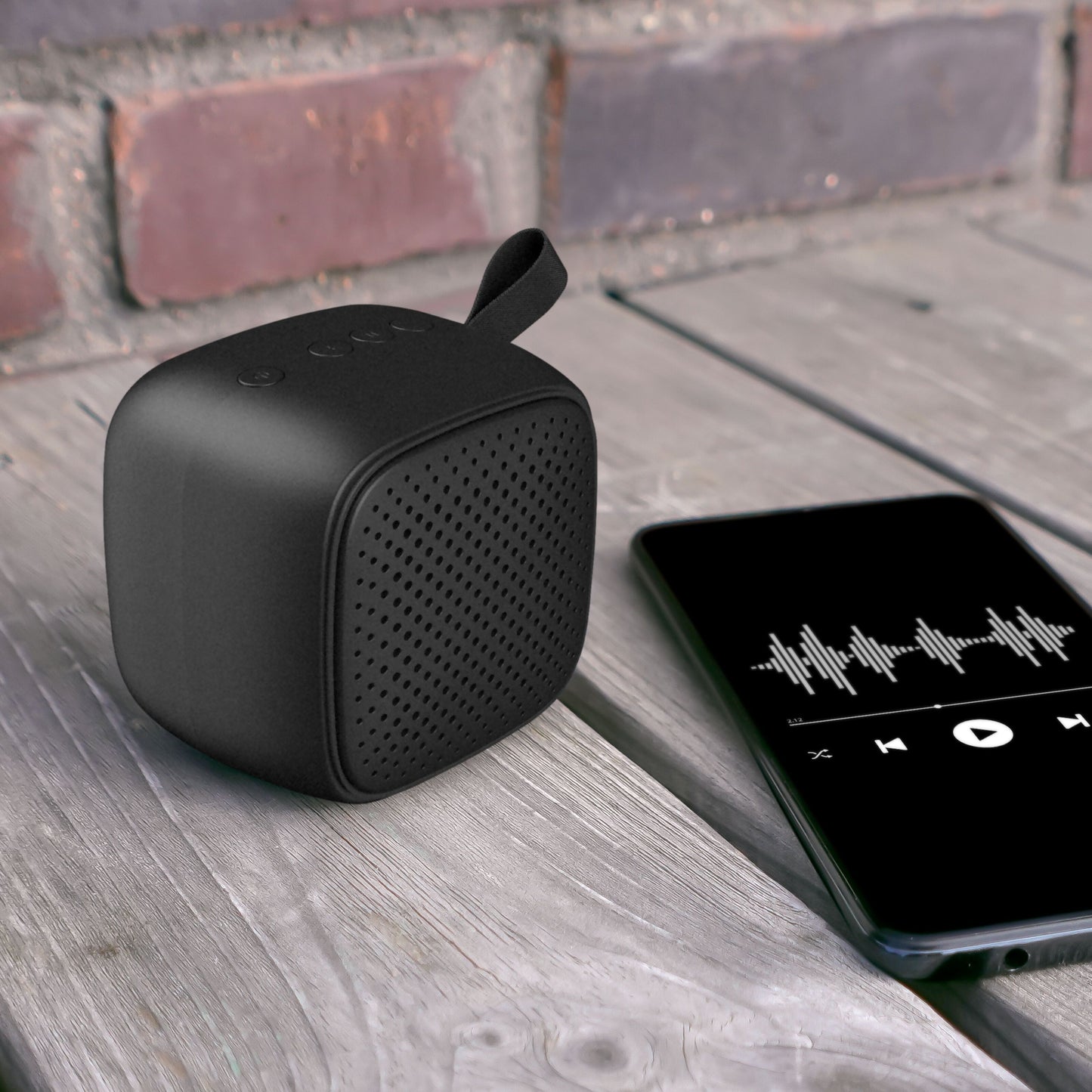 Linocell Trådløs højttaler Sort Bluetooth med vævet rem, set på træ ved siden af smartphone med musikbølge display.