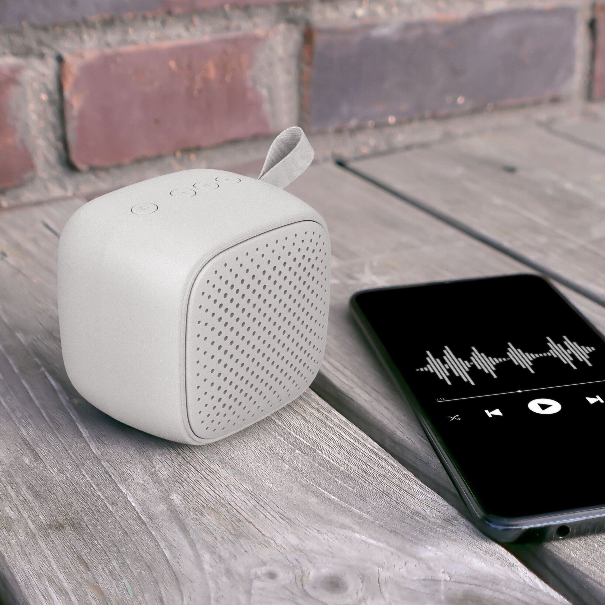 Linocell trådløs højttaler grå ved siden af smartphone med musikapp, placeret på træbord, synlig murstensvæg.