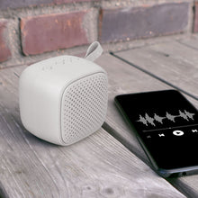 Linocell trådløs højttaler grå ved siden af smartphone med musikapp, placeret på træbord, synlig murstensvæg.