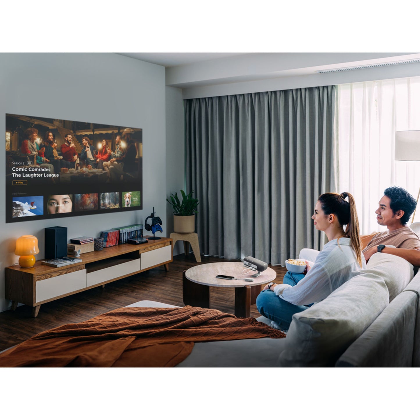 ViewSonic M1XG bærbar projektor med Harman Kardon-højttalere projicerer i stue, træbord og fjernbetjening synlig.