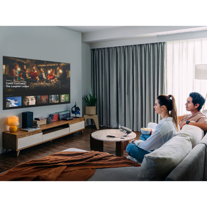 ViewSonic M1XG bærbar projektor med Harman Kardon-højttalere projicerer i stue, træbord og fjernbetjening synlig.
