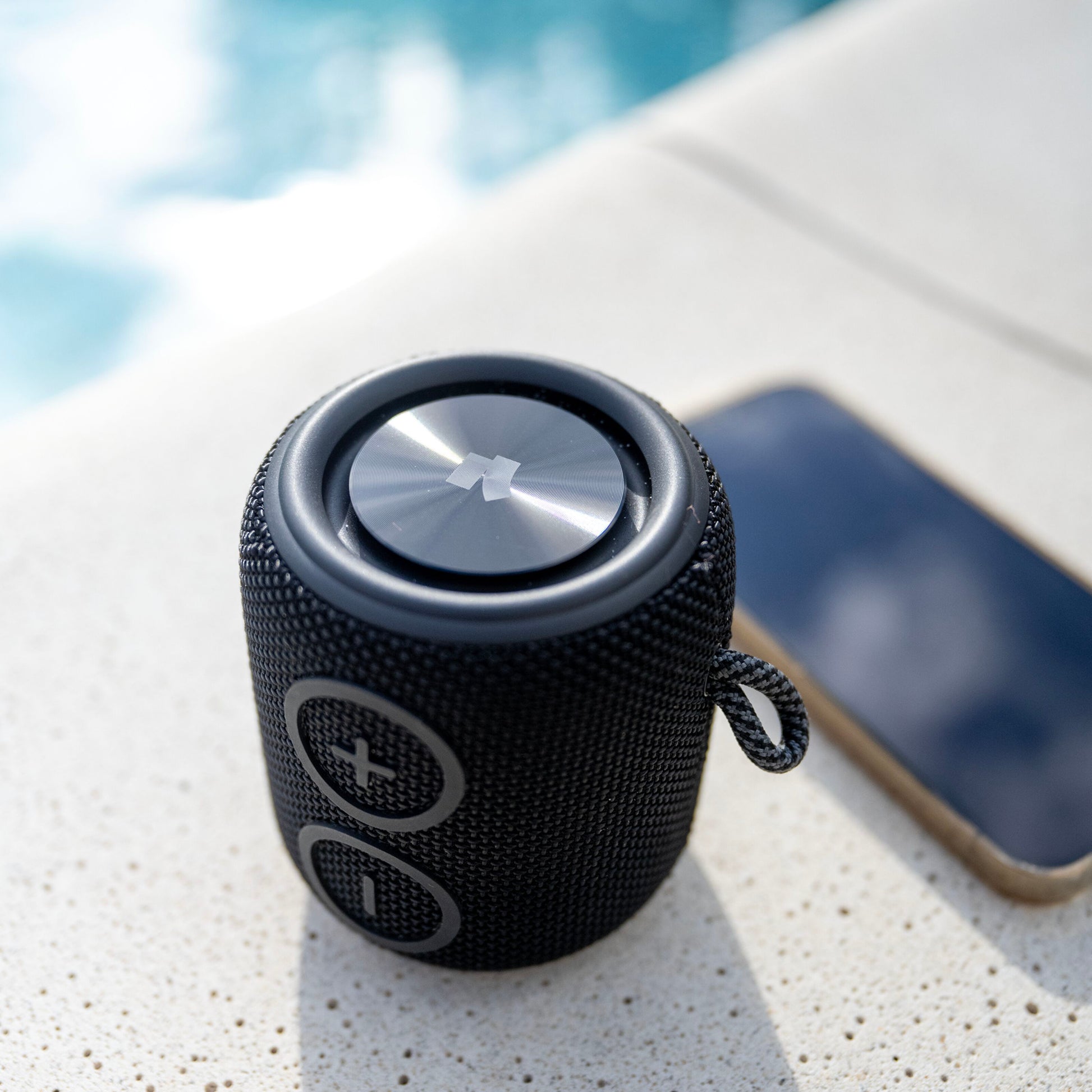 Nomadelic Loud 705 vandtæt Bluetooth 5.0 højttaler sort, store plus og minus-knapper, ved siden af smartphone ved pool.