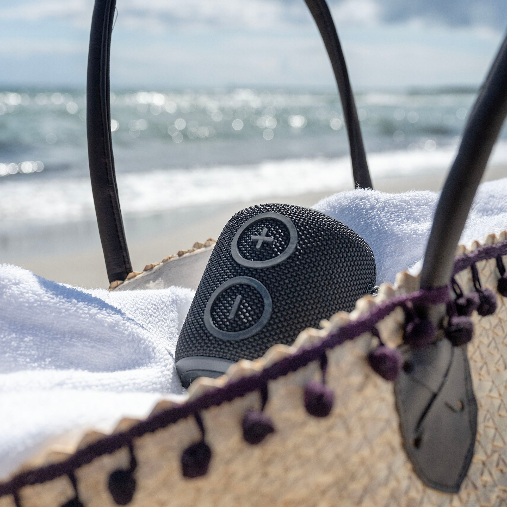 Nomadelic Loud 705 vandtæt Bluetooth-højttaler sort på hvidt håndklæde i strå strandtaske, uskarp strand i baggrunden.