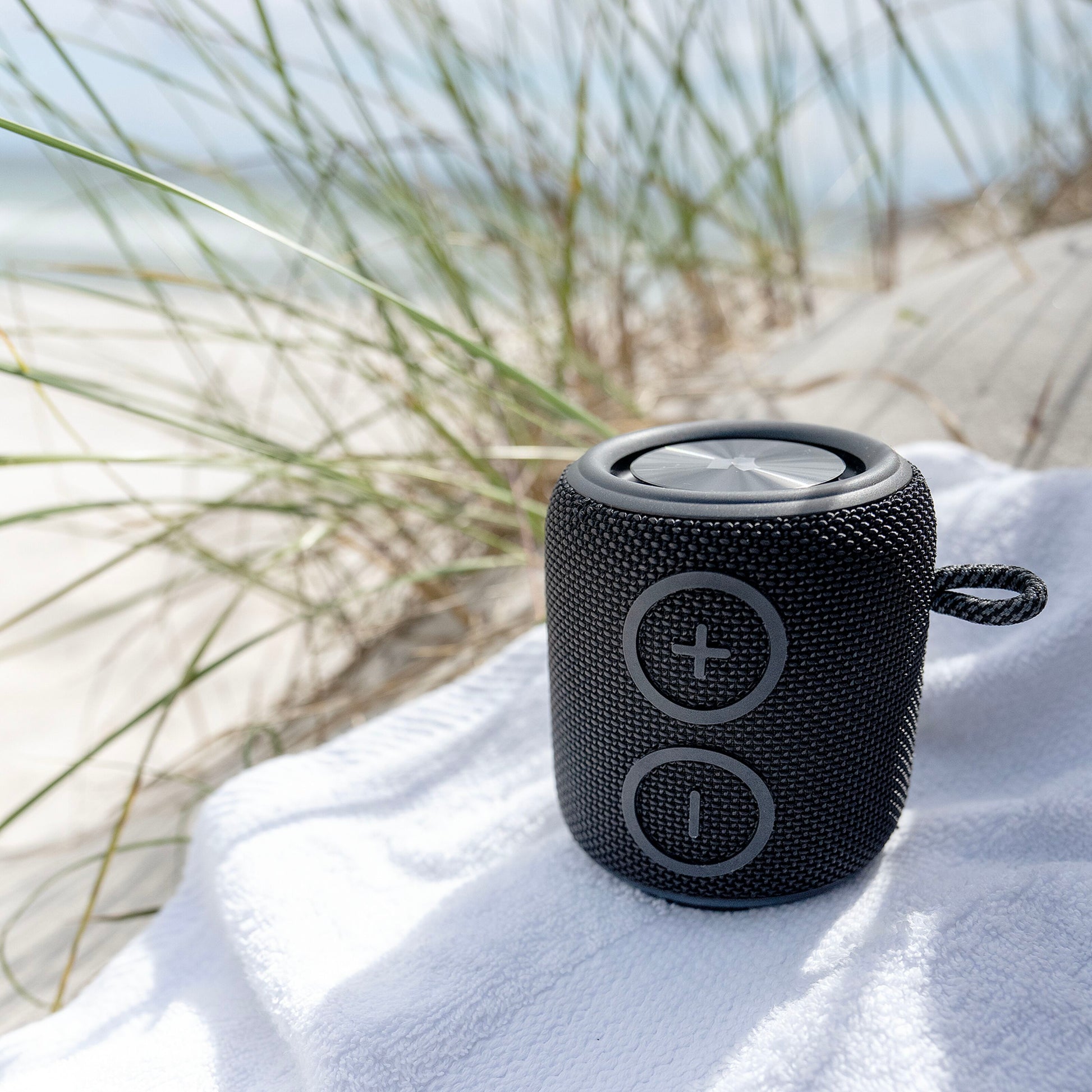 Nomadelic Loud 705 Bluetooth-højttaler sort med store volumeknapper, Bluetooth 5.0 og IPX7 vandtæt, ude på håndklæde.