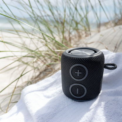 Nomadelic Loud 705 Bluetooth-højttaler sort med store volumeknapper, Bluetooth 5.0 og IPX7 vandtæt, ude på håndklæde.