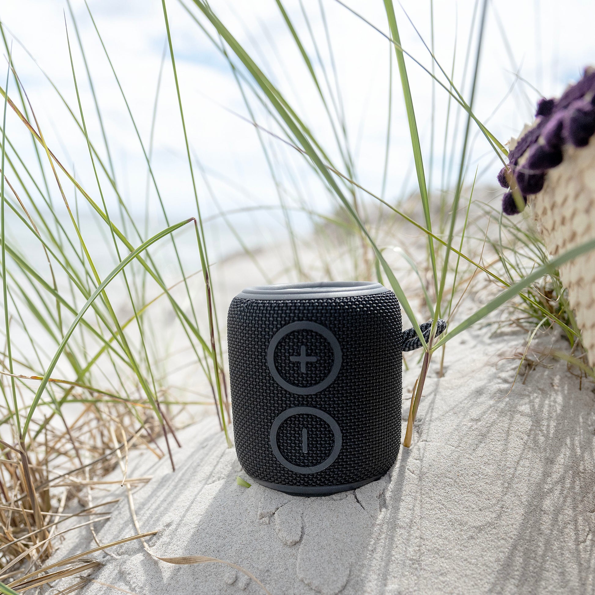 Nomadelic Loud 705 sort vandtæt Bluetooth-højttaler med stofbetræk på klit, hav og himmel sløret i baggrunden.