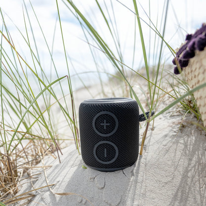 Nomadelic Loud 705 sort vandtæt Bluetooth-højttaler med stofbetræk på klit, hav og himmel sløret i baggrunden.
