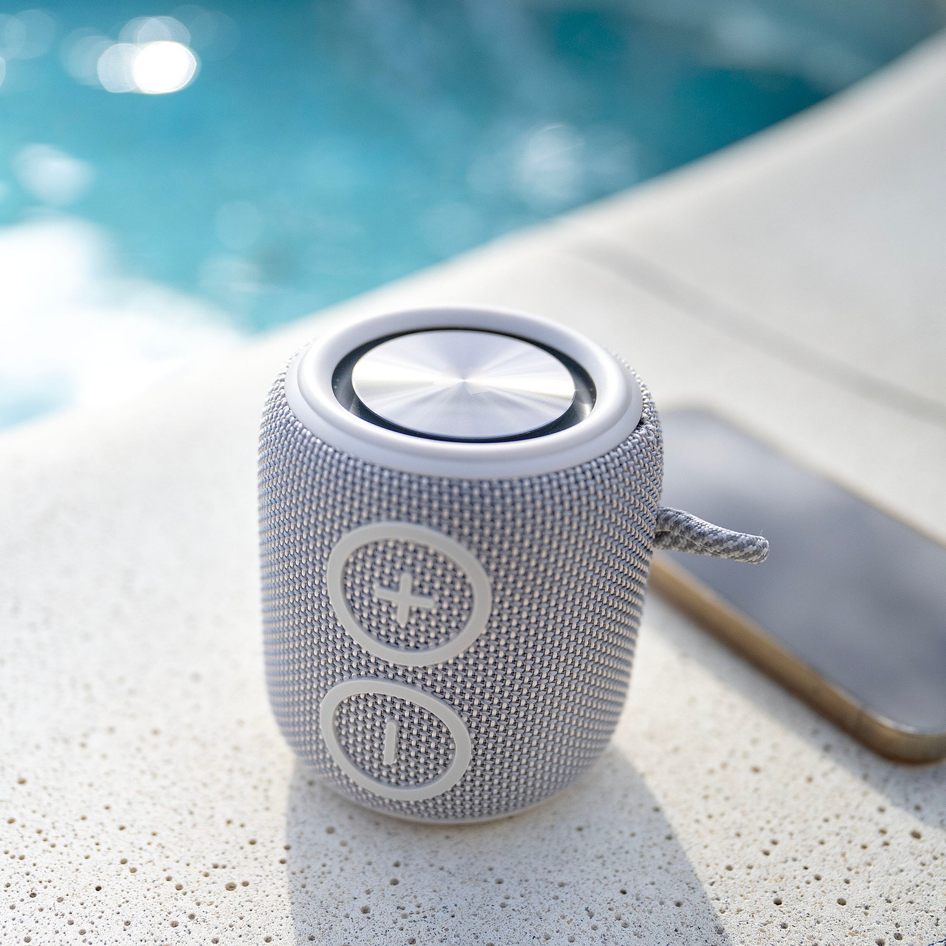 Nomadelic Loud 705 vandtæt Bluetooth-højttaler grå med store volumeknapper ved siden af smartphone nær swimmingpool.