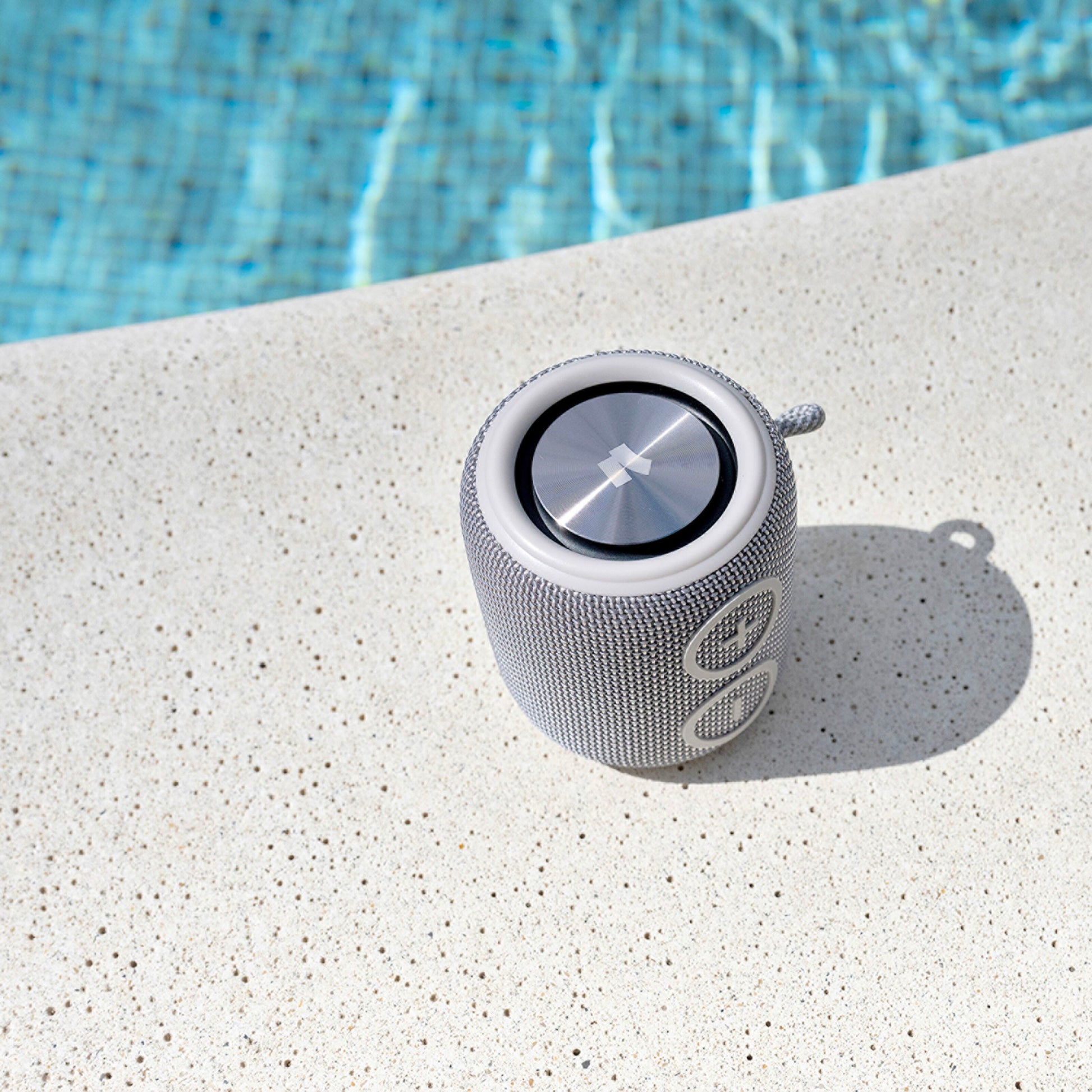 Nomadelic Loud 705 vandtæt Bluetooth-højttaler sort på tekstureret poolkant med blåt vand i baggrunden.