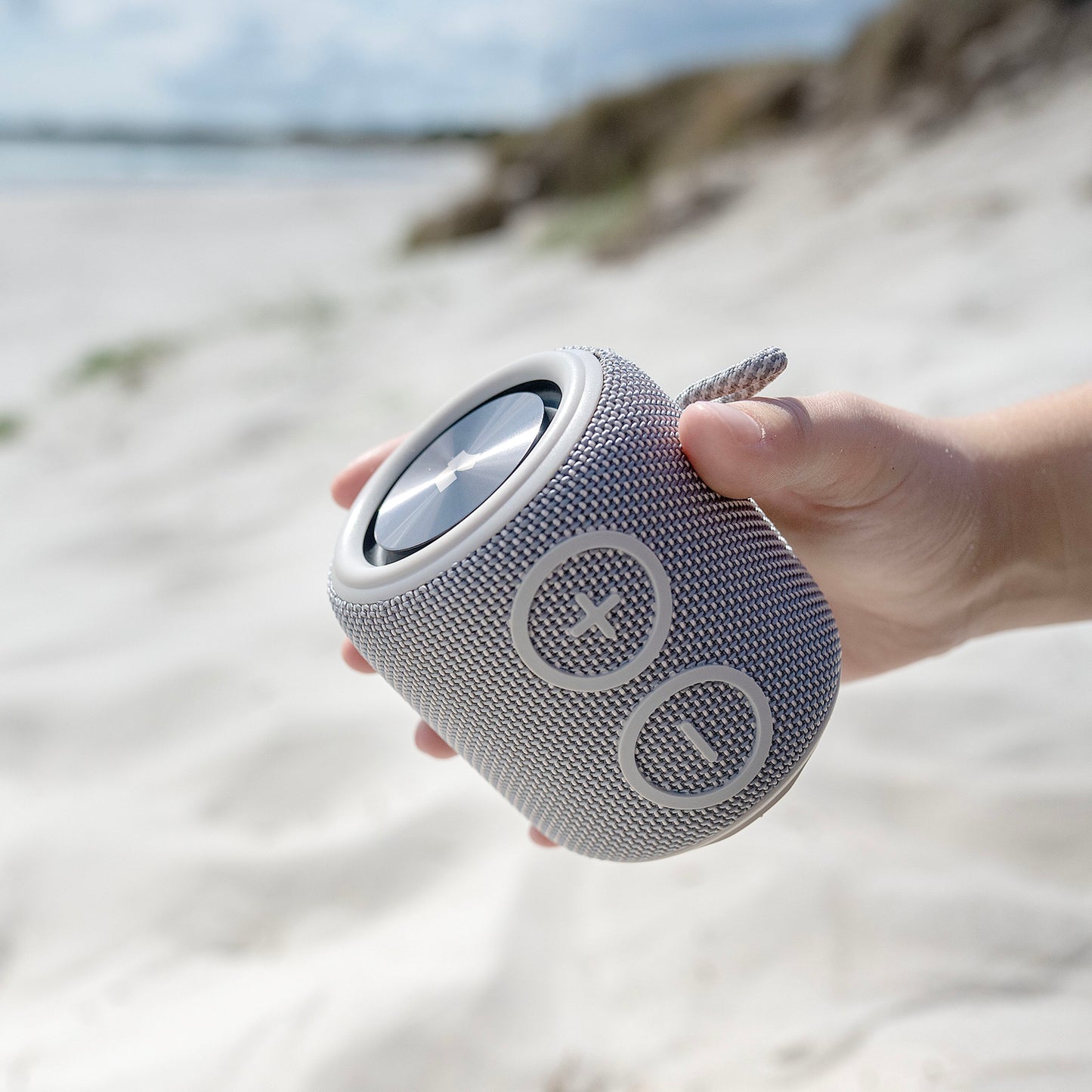 Nomadelic Loud 705 grå vandtæt Bluetooth-højttaler med store plus- og minus-volumenknapper ses i hånd ved strand.