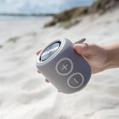 Nomadelic Loud 705 grå vandtæt Bluetooth-højttaler med store plus- og minus-volumenknapper ses i hånd ved strand.