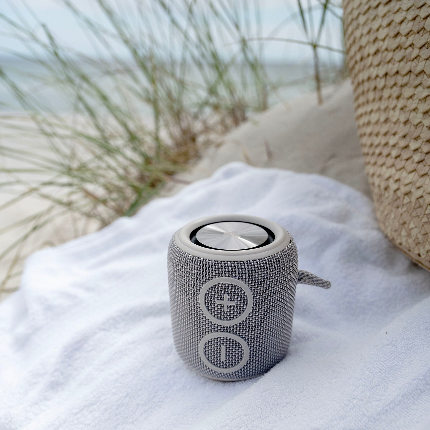 Nomadelic Loud 705 vandtæt Bluetooth-højttaler grå på hvidt håndklæde ved strand med klitgræs og flettet taske.