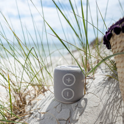 Nomadelic Loud 705 vandtæt Bluetooth-højttaler grå på hvidt sand med strandgræs og flettet taske ved siden af.
