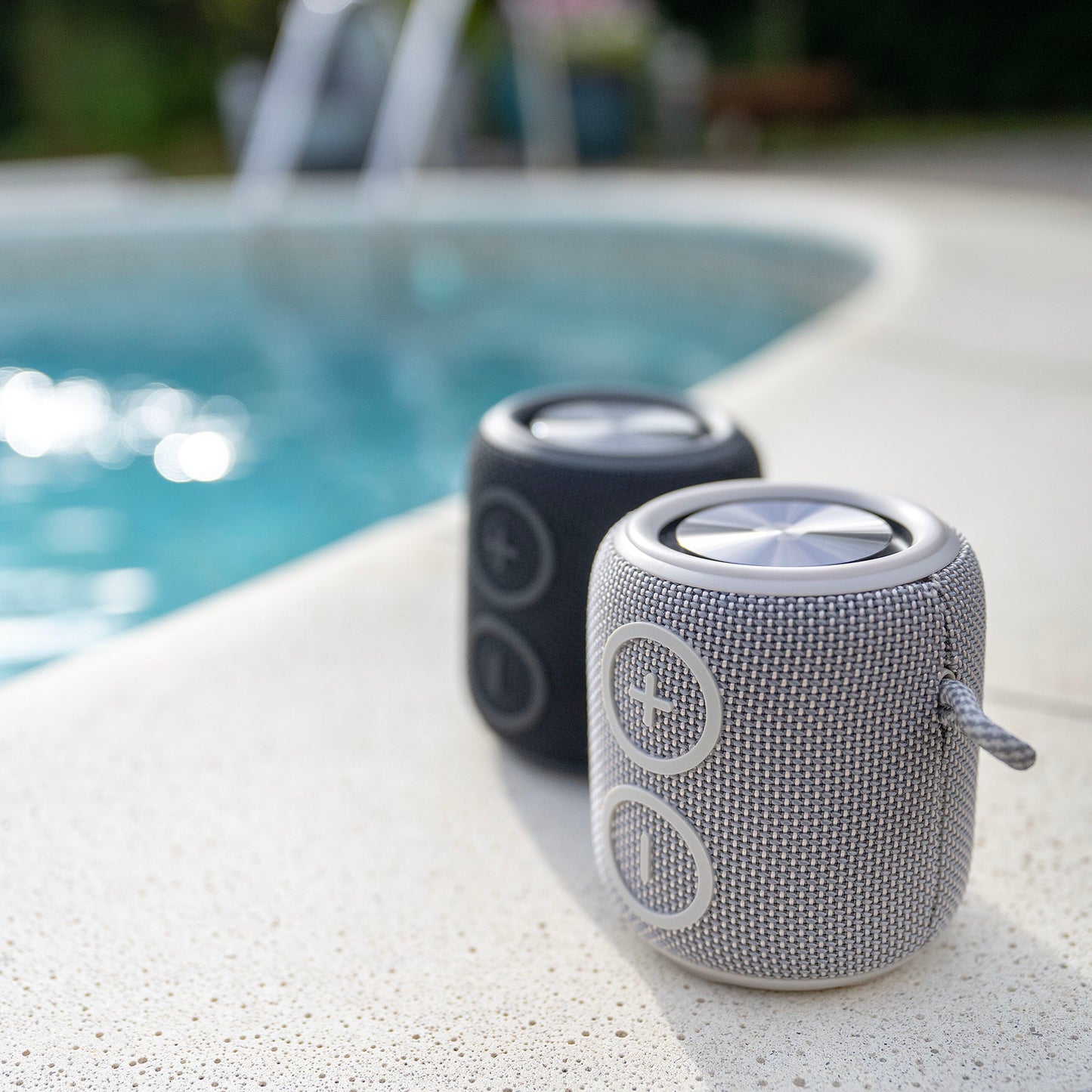 Nomadelic Loud 705 vandtæt Bluetooth-højttaler grå foran og sorter bagved ved poolkant, sløret pool i baggrunden.