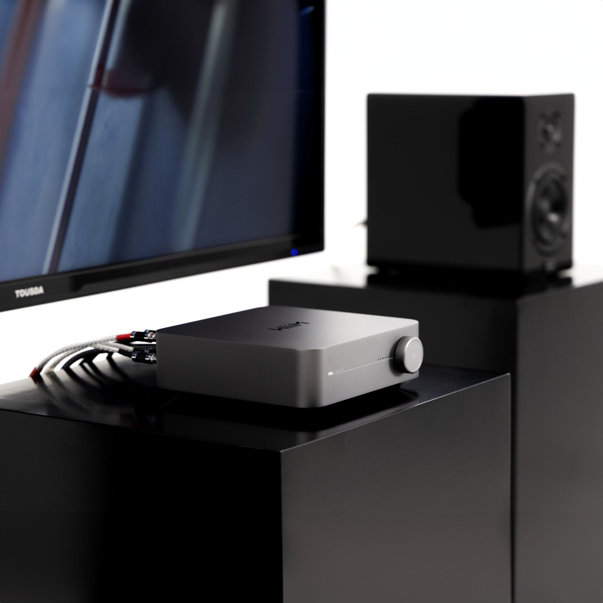 Wiim Amp Stereo sølv forstærker med streaming, synlige kabler, ved siden af fladskærms-tv og sort højttaler.