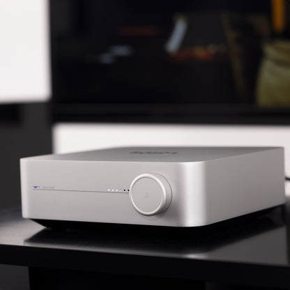 Wiim sølv forstærker med streaming fra Amp Stereo, LED-lys og trekantet knap på front, set fra siden på sort bord.