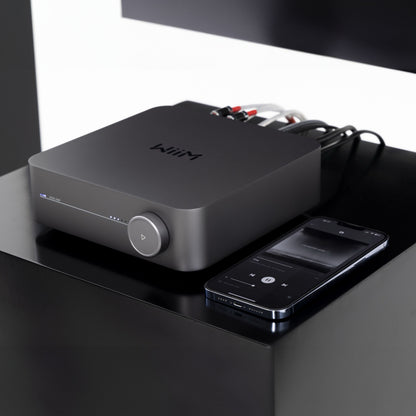 WiiM forstærker med streaming, model Amp Stereo, sort, tilsluttet lydkabler, synlig smartphone med musikafspiller-app.