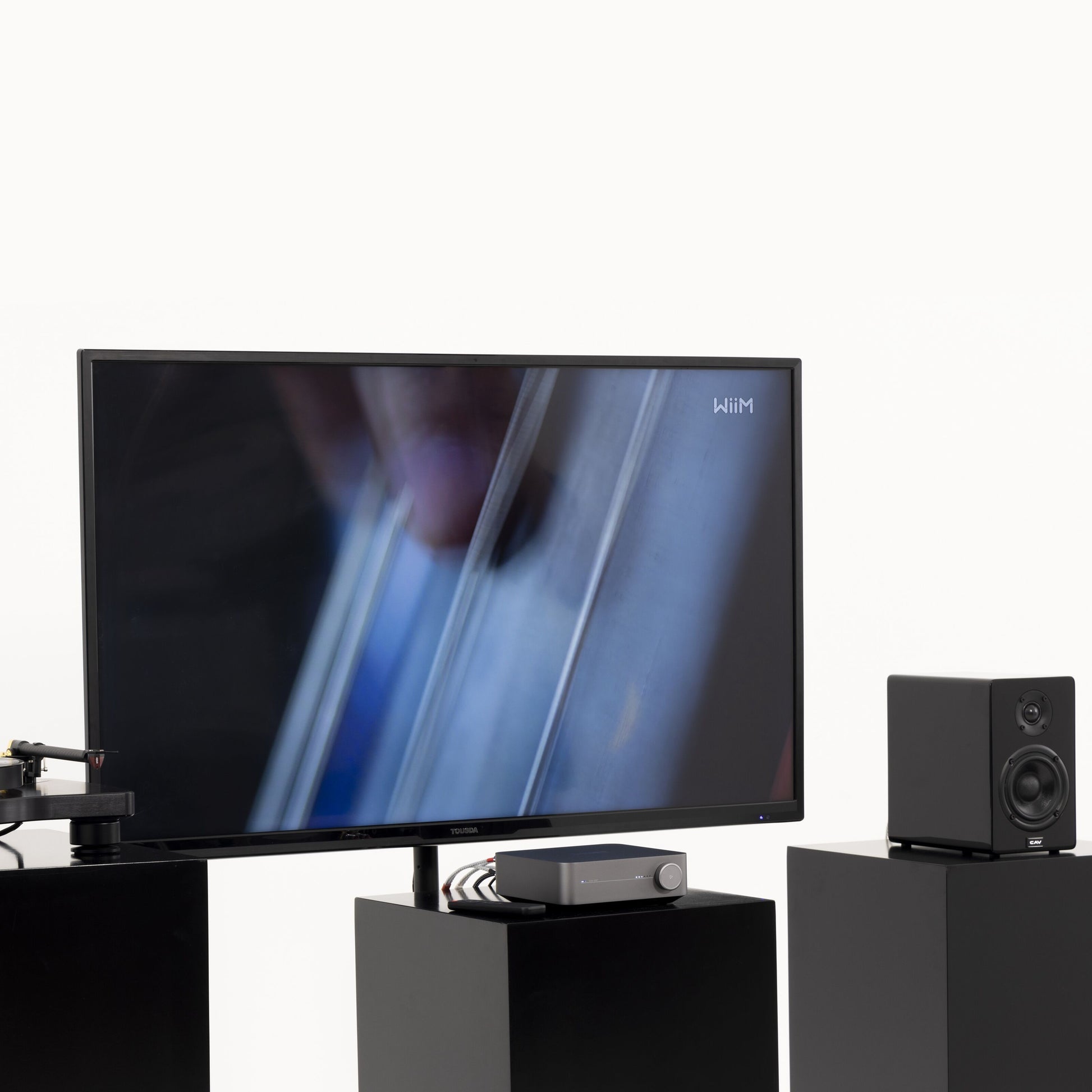 Wiim forstærker med streaming via Amp Stereo, fladskærms-tv, boghylde-højttaler og pladespiller på sorte stativer.
