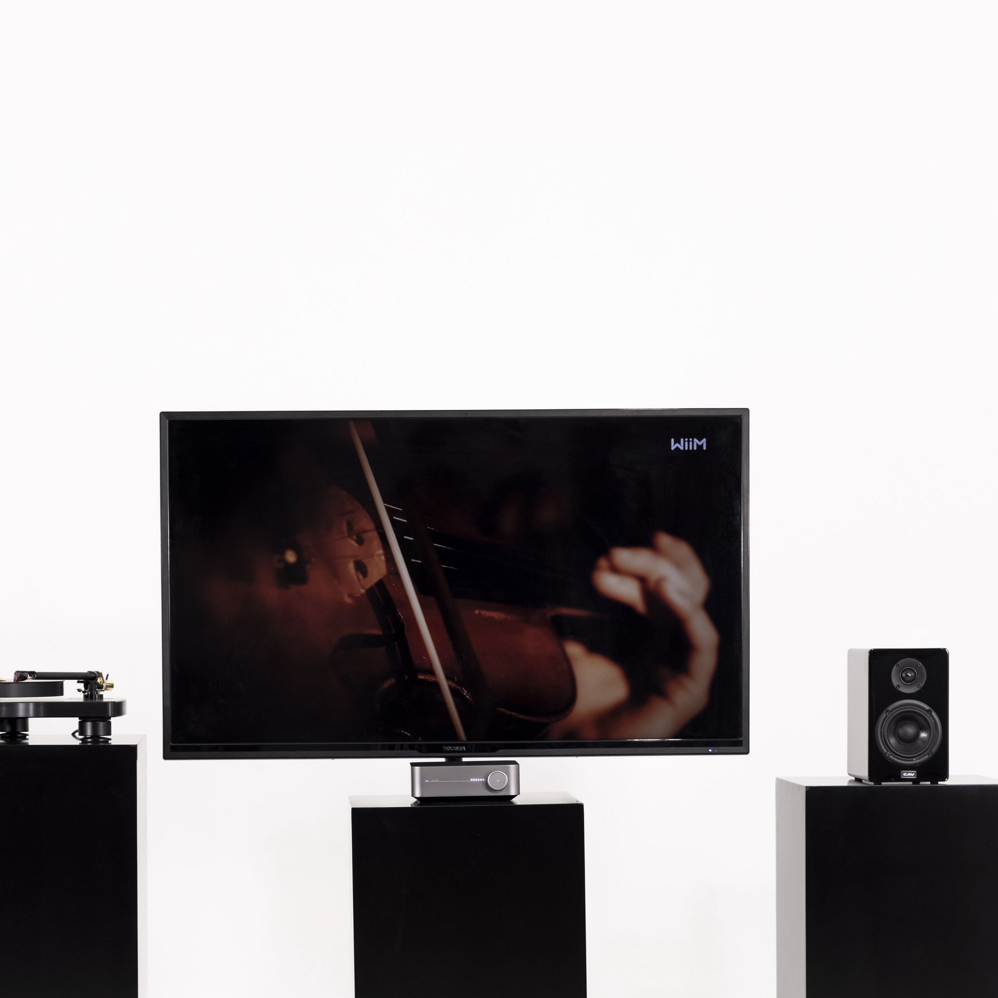 Fladskærms-TV på sort stander med hænder der spiller violin, to sorte højttalere og pladespiller, Wiim Forstærker Amp Stereo.
