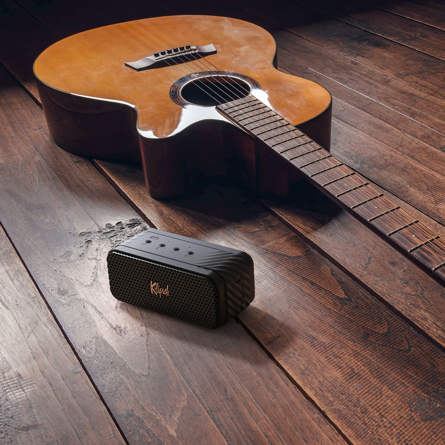 Brun akustisk guitar ved siden af sort Klipsch Nashville Bluetooth-højttaler på trægulv, let refleksion på guitar.