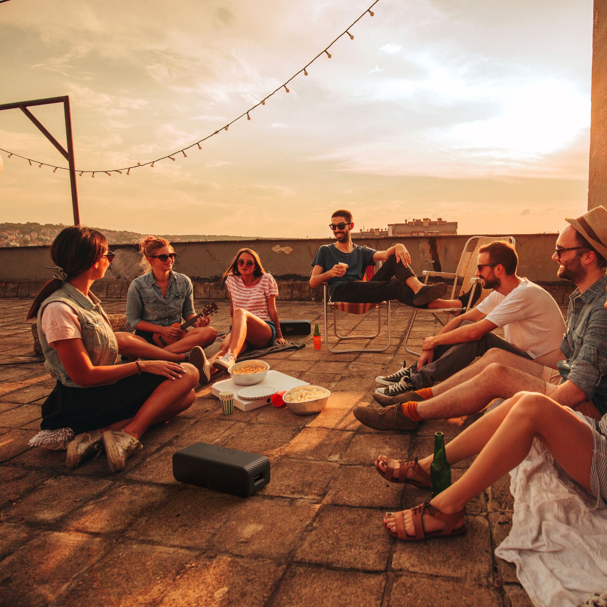 Klipsch Detroit Bluetooth-højttaler sorterer sine i brug på tagterrasse omgivet af unge voksne, drikkevarer og snacks.