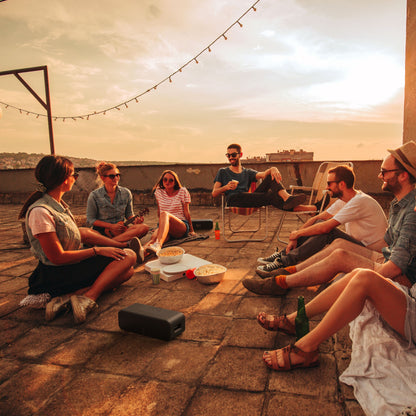 Klipsch Detroit Bluetooth-højttaler sorterer sine i brug på tagterrasse omgivet af unge voksne, drikkevarer og snacks.