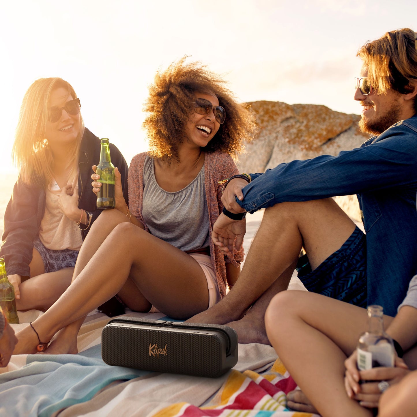 Klipsch Detroit Bluetooth-højttaler sort, placeret foran gruppe på strand med klipper og solnedgang i baggrunden.