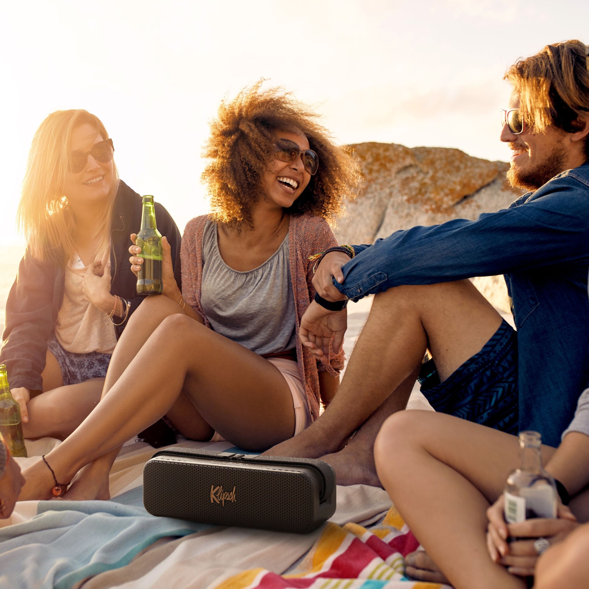 Klipsch Detroit Bluetooth-højttaler sort, placeret foran gruppe på strand med klipper og solnedgang i baggrunden.