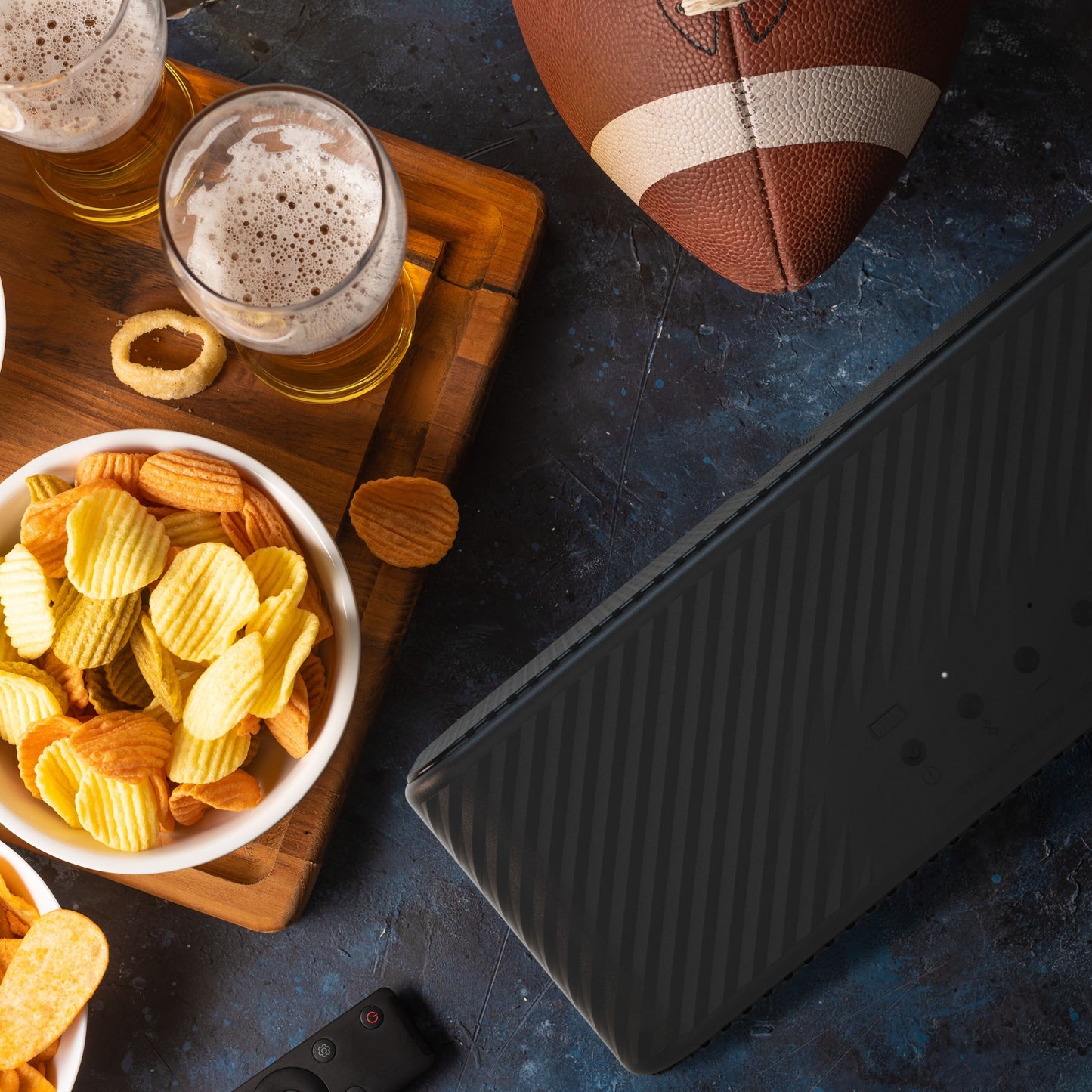 Klipsch Detroit Bluetooth-højttaler sort, 20+ timer batteritid, topview med fodbold, snacks og øl på mørk overflade.