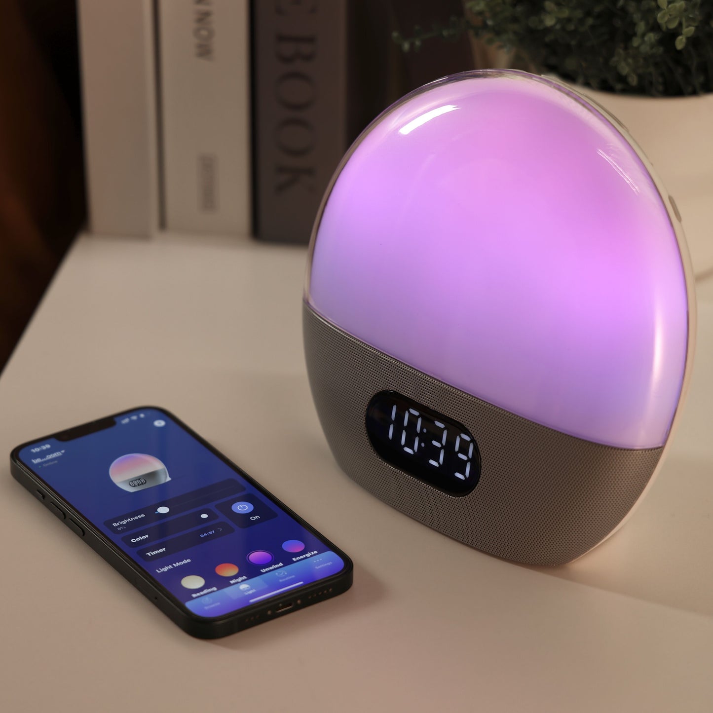 WiiM Vækkeur wake-up light med lilla lys på skrivebord, smartphone med lysstyring-app ved siden af, bøger i baggrunden.