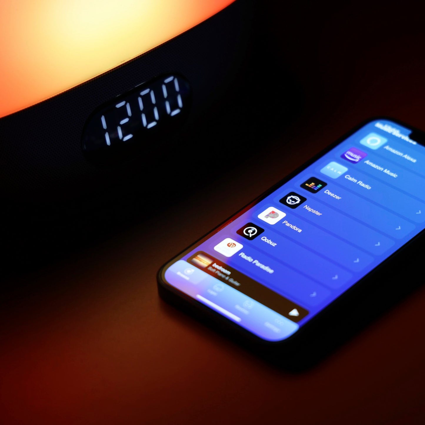WiiM vækkeur med wake-up light viser 12:00, varm orange lys, smartphone med musikapps ses ved siden af.