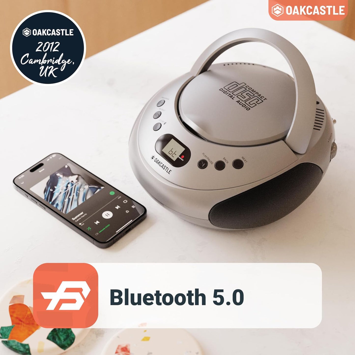 Oakcastle CD250 bærbar CD-afspiller med Bluetooth ved siden af smartphone, Oakcastle-logo og tekst "2012 Cambridge, UK.