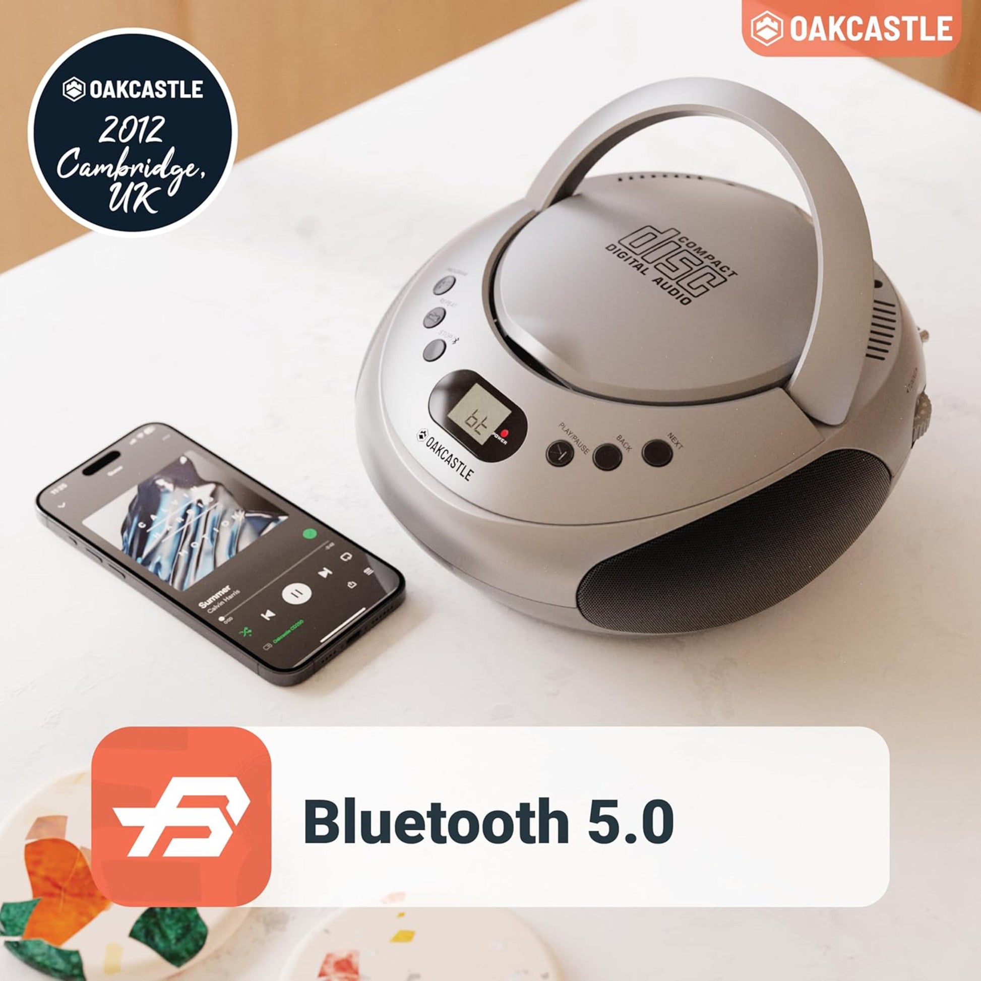 Oakcastle CD250 bærbar CD-afspiller med Bluetooth ved siden af smartphone, Oakcastle-logo og tekst "2012 Cambridge, UK.