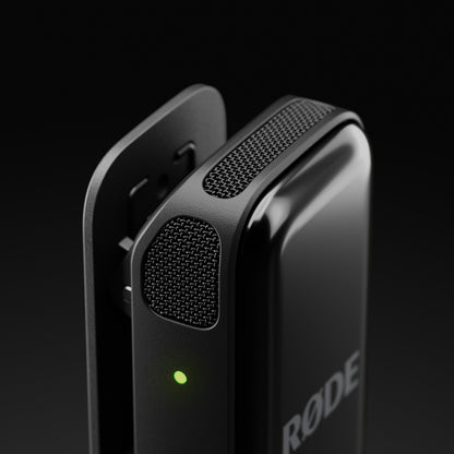 Røde Trådløs mikro lyn transmitter i sort med clip, mesh gitter og grøn indikatorlys mod mørk sløret baggrund.